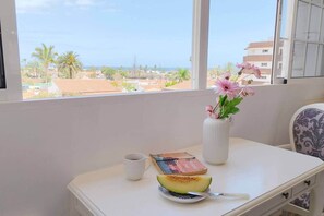 Ein Zimmer mit Fensterblick auf einen Strand, ein weißer Tisch mit einem Buch, eine Vase mit Blumen und ein Teller mit einem Melonenscheibchen.