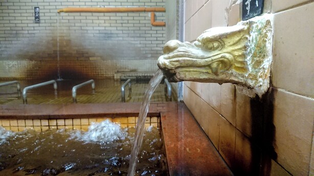 A water fountain with a decorative head, a tiled wall, and metal handrails in the background. - Hotel Yuou Onsen  (Kofu)