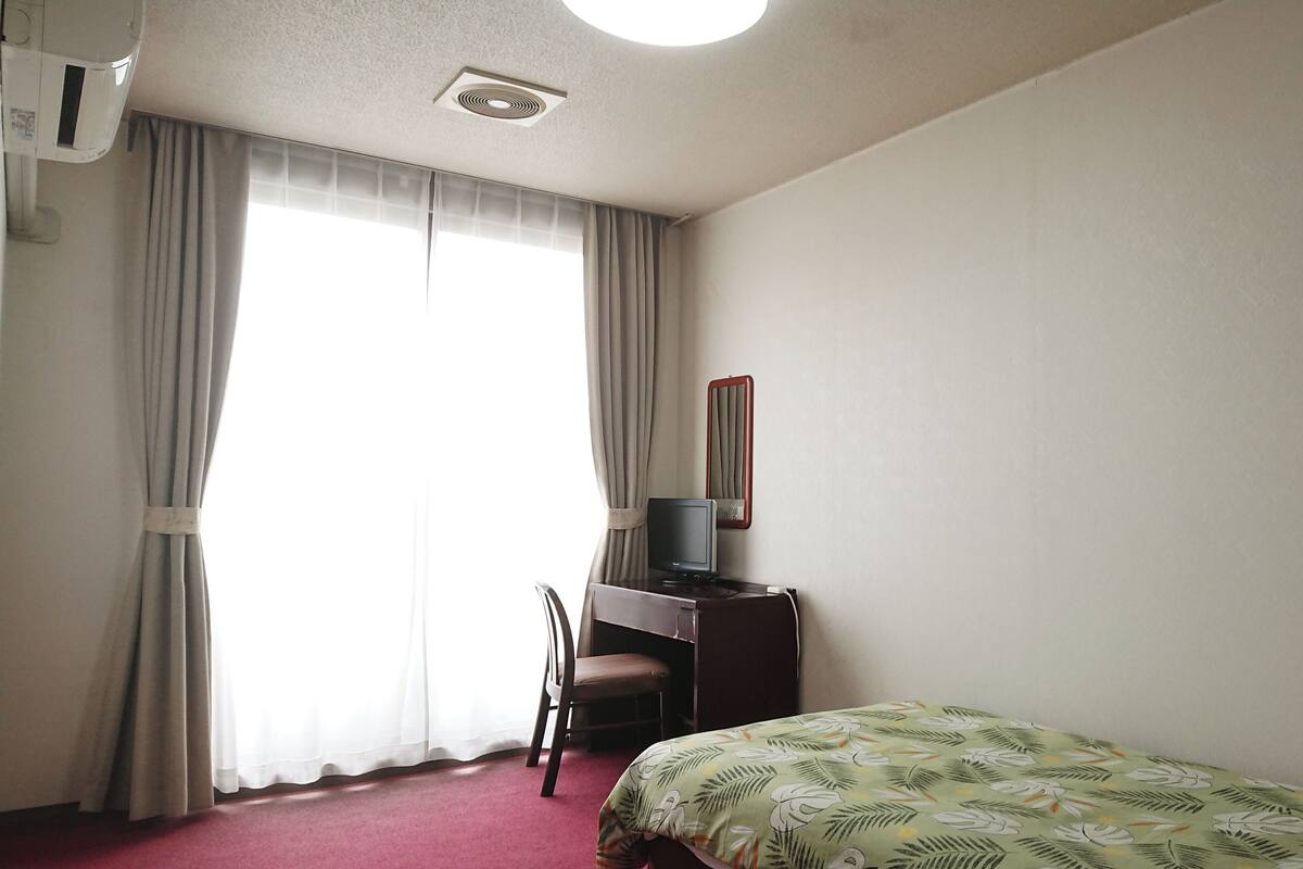 A hotel room with a bed, a desk with a chair, a television, and a window with curtains.