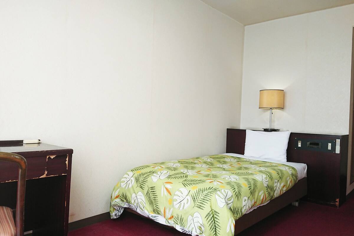 A hotel room with a single bed, a desk, a chair, a lamp, and a mirror.