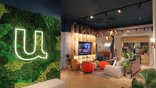 A modern interior with a green wall, a large illuminated 'U', a TV, and a seating area with orange and white chairs. - Student Only Bargate House (Southampton)
