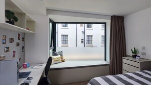 A bedroom with a large window, a desk with a laptop and keyboard, a bed with striped bedding, a small bedside table with a plant, and a shelf with photos and a teddy bear. - Student Only Bargate House (Southampton)
