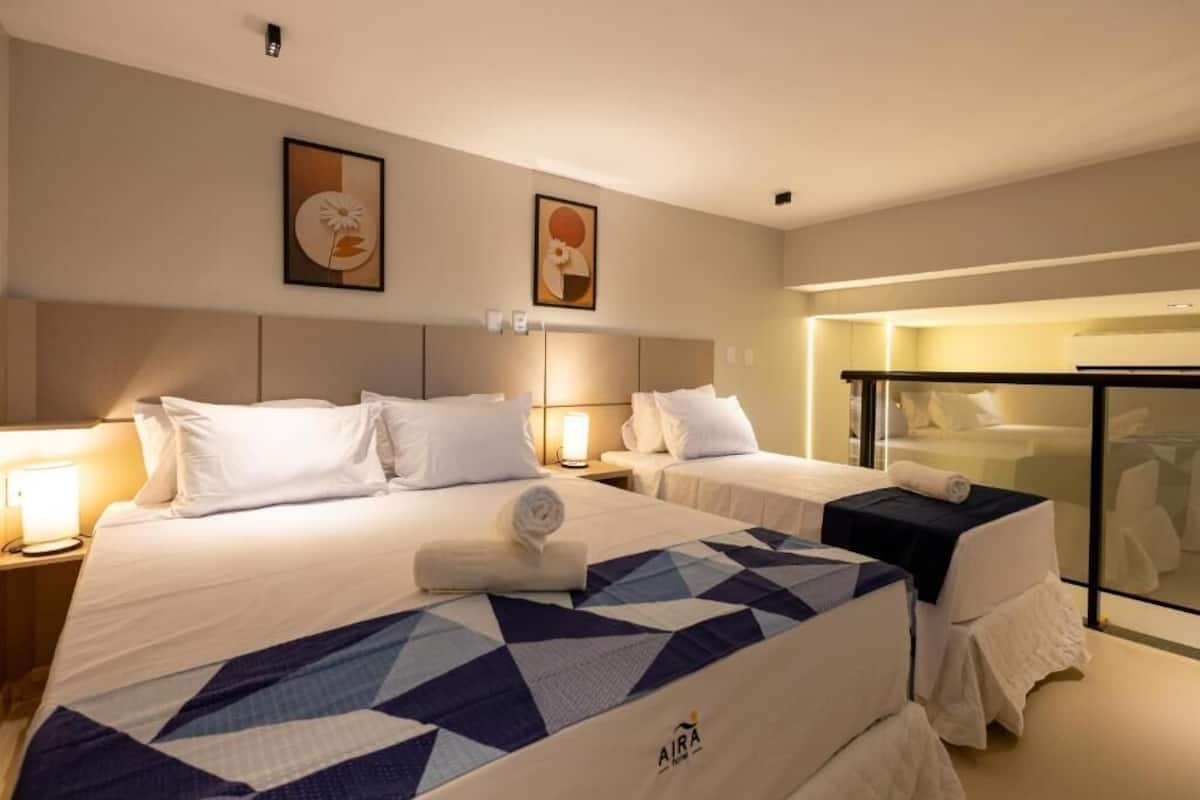 Ett hotellrum med två sängar, en stor säng med ett geometriskt mönster på sängöverkastet och en mindre säng med ett enfärgat sängöverkast. Det finns väggmonterade lampor, en tavla och en glasvägg med en handdukshängare.