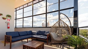 A modern living room with a blue sectional sofa, a wooden coffee table, a hanging chair, and a large window offering a city view. - Aira Hotel (Sobral)