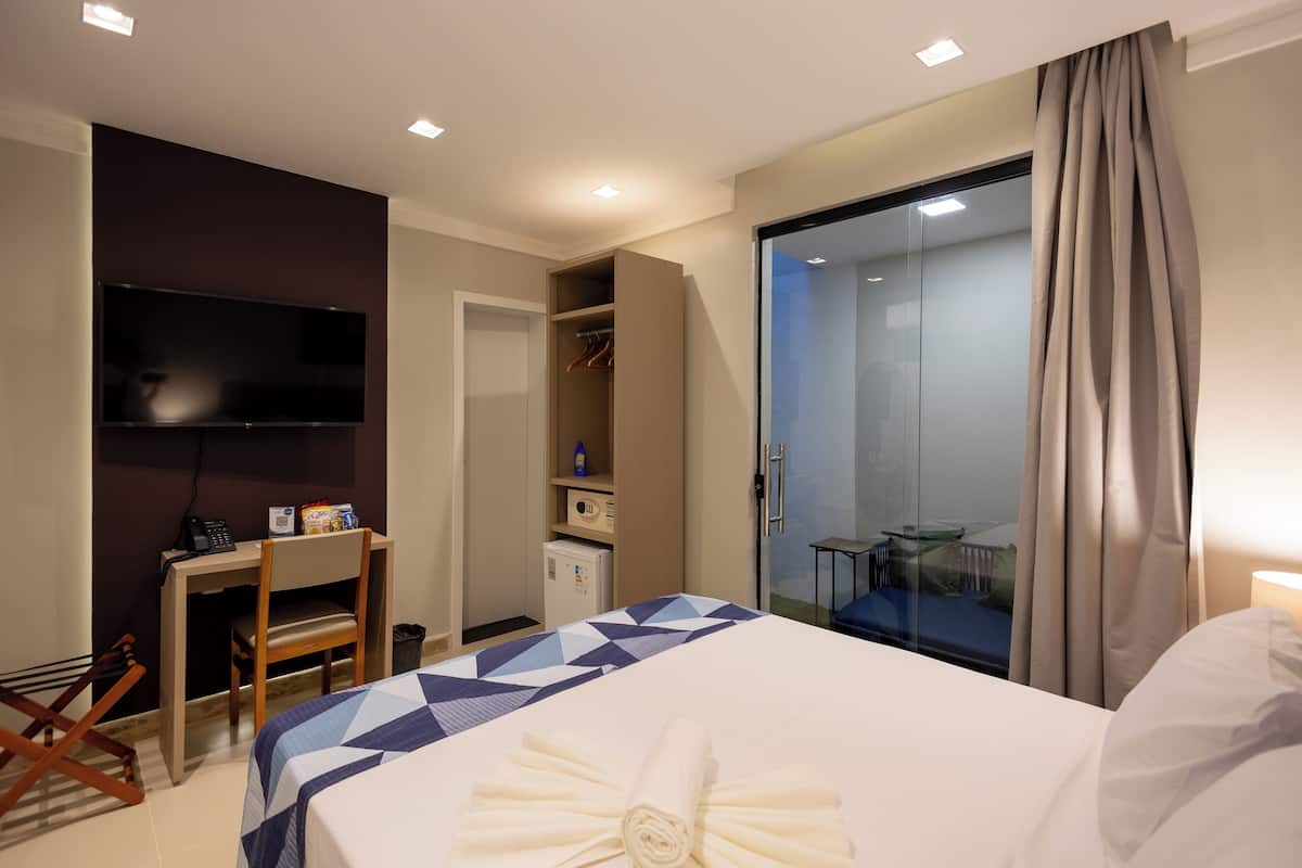 Ett hotellrum med en säng, ett skrivbord, en stol, en TV, en garderob och en glasdörr som leder ut till en balkong.