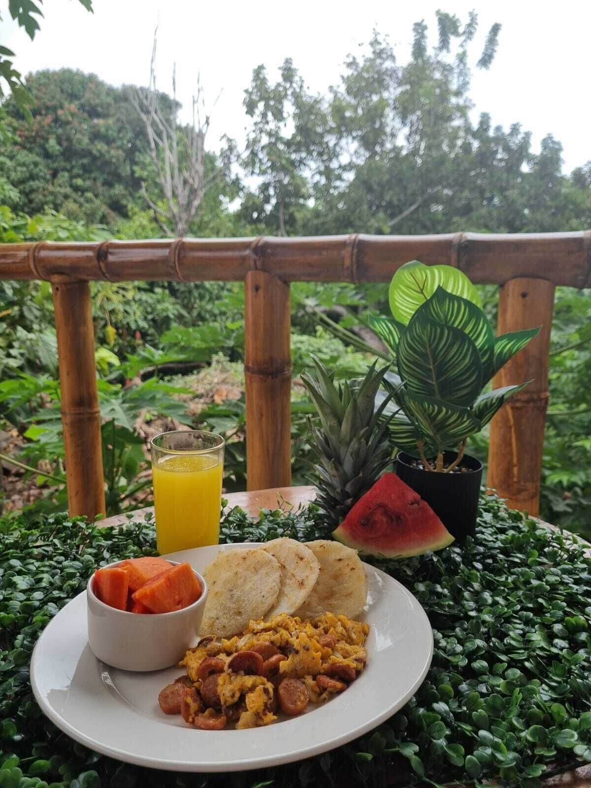 A breakfast plate with toast, scrambled eggs, sausage, and a bowl of fruit, served outdoors with a view of lush greenery and a bamboo fence.
