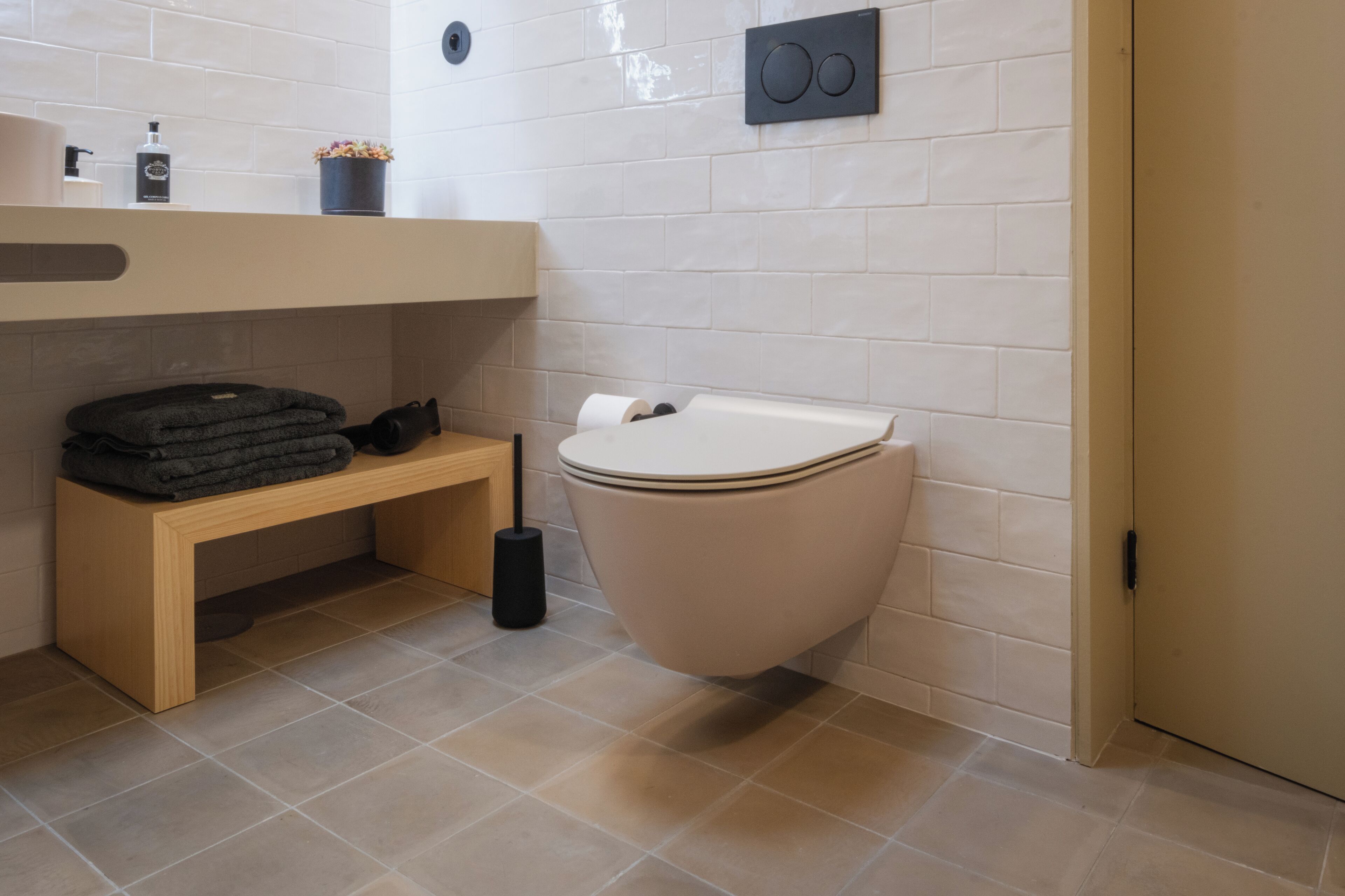 A modern bathroom with a toilet, a bench, a sink, and a small shelf with toiletries.