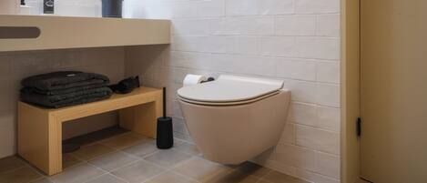 A modern bathroom with a toilet, a bench, a sink, and a small shelf with toiletries.