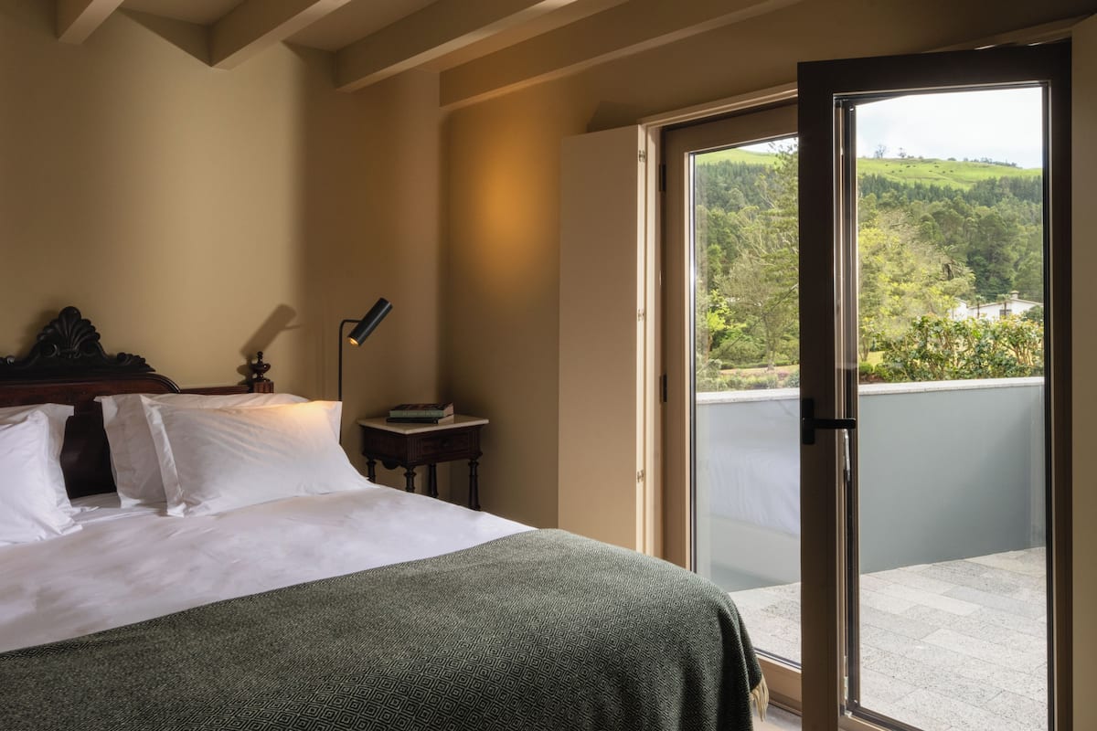 Een slaapkamer met een bed, een nachtkastje met een lamp, en uitzicht op een balkon met een mooi landschap.
