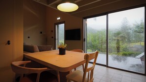 A dining area with a wooden table, chairs, a potted plant, and a large window with a view of greenery. - Casa Nas Caldeiras (Povoacao)