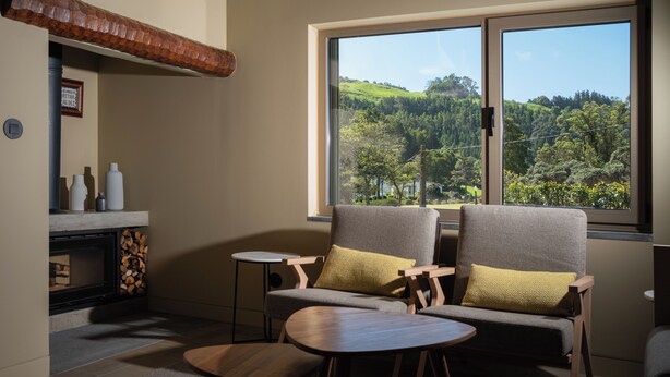 A modern living room with a fireplace, two sofas with cushions, a small table, and a large window offering a view of a forested hillside. - Casa Nas Caldeiras (Povoacao)