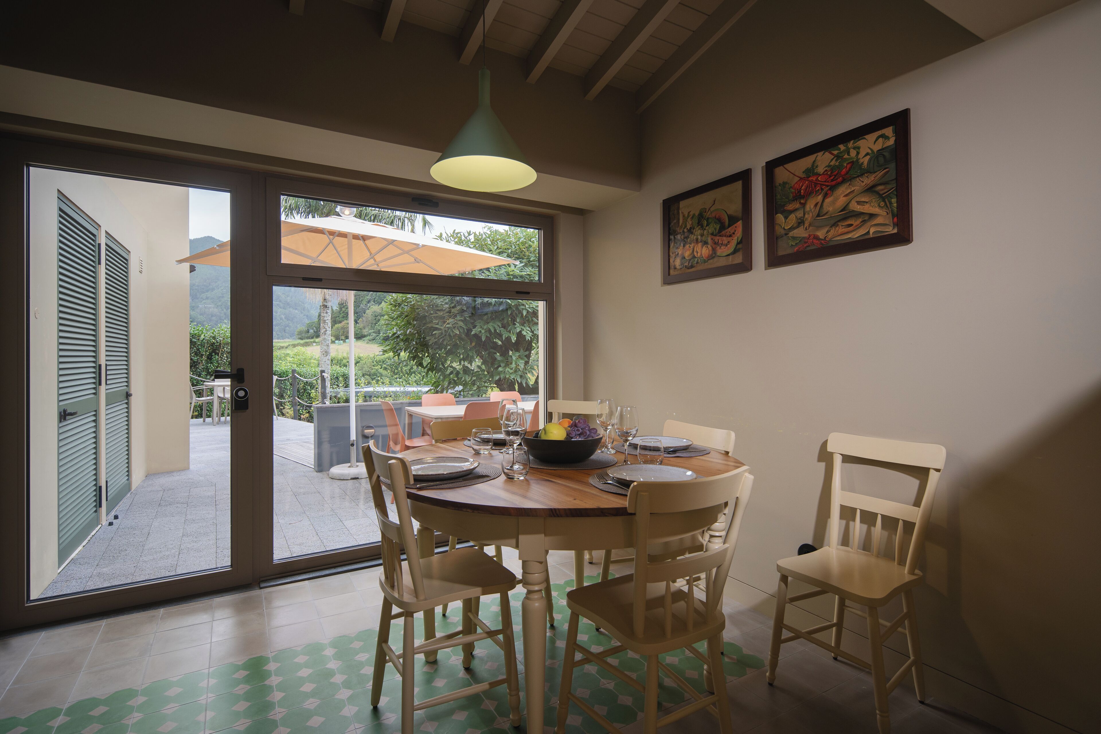 A dining area with a round table, four chairs, a bowl of fruit, and a window with an umbrella outside.