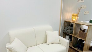 A white leather sofa with two cushions, a wooden shelf with rolled towels, and a small round table.