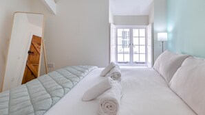A hotel room with a bed, a mirror, a window with blinds, and a nightstand with a lamp.