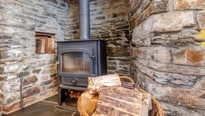 A stone-walled room with a wood-burning stove, a basket of firewood, and a small wooden shelf.