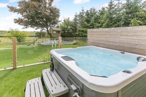 A hot tub with a wooden deck and a view of a playground in the background.