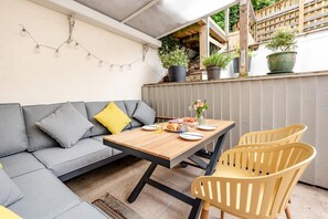 A patio with a wooden table, two chairs, a grey sofa with cushions, and potted plants.