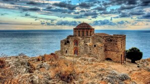 An ancient stone building with a dome and arched windows, situated on a rocky coastline with a view of the sea.