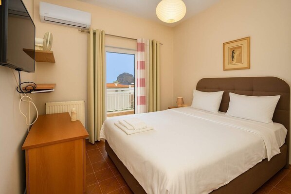 A hotel room with a bed, a desk, an air conditioning unit, a window with curtains, and a framed picture on the wall. - Cosmo Room 2 | Seaside Retreat | Monemvasia Castle (Monemvasia)