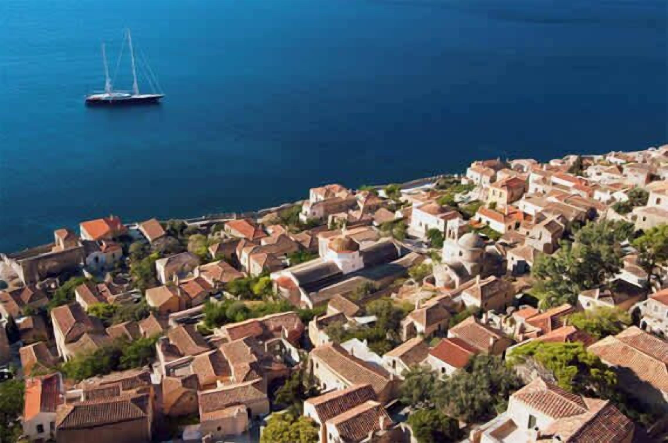 Un pueblo costero con edificios muy juntos, un velero en el agua y un paisaje montañoso al fondo.