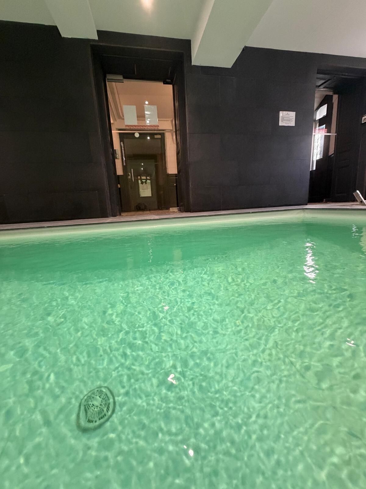 Ein Innenpool mit klarem Wasser, umgeben von dunklen Wänden und einer sichtbaren Tür zu einem anderen Raum.
