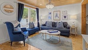Una sala de estar con un sillón azul, un sofá, una mesa de centro redonda y una araña.
