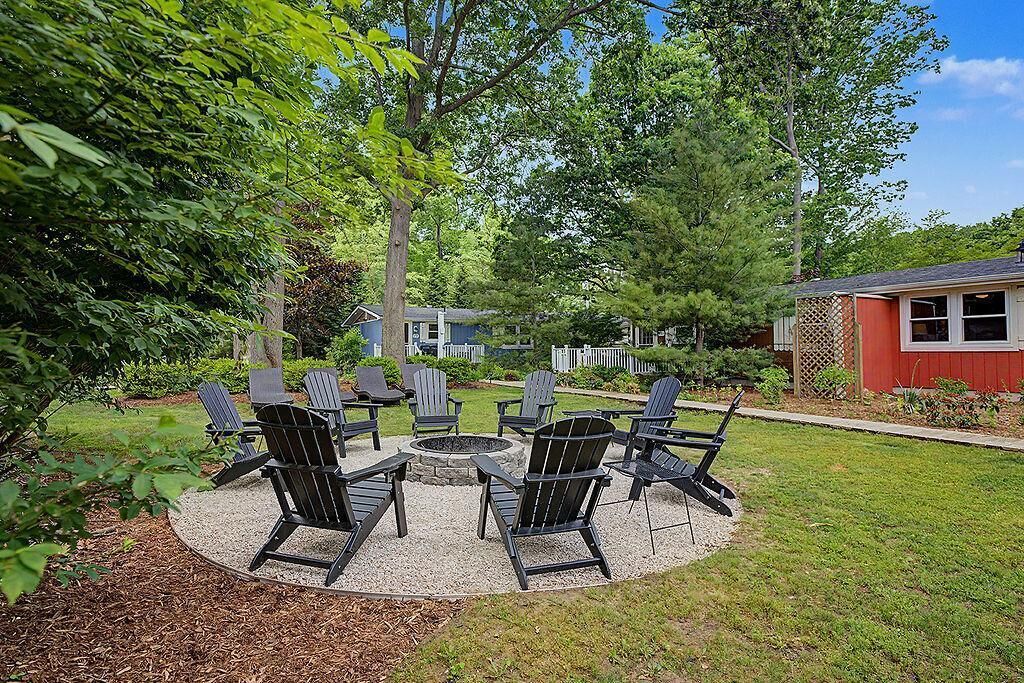 A circular outdoor seating area with Adirondack chairs and a fire pit, surrounded by a gravel circle, with a red building and trees in the background.