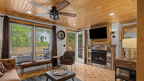 A cozy living room with a fireplace, a ceiling fan, and a large window overlooking a deck.