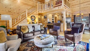 A spacious log cabin interior with a living area featuring leather sofas, a coffee table, and a large rug. A wooden staircase leads to an upper level with a balcony. - Timber Nest Goshorn lake dock, pool, town HOT TUB! (Saugatuck)