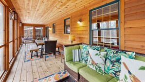 A wooden cabin interior with a wooden deck, a green sofa with tropical pillows, a dining table with chairs, and large windows. - Timber Nest Goshorn lake dock, pool, town HOT TUB! (Saugatuck)