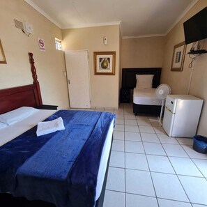 A hotel room with a bed, a desk, a chair, a small refrigerator, a fan, and a door.