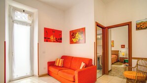 A living room with a red sofa, two wall-mounted paintings, a wicker chair, and a view into a bedroom through an open door. - KNOWING ROME. (Roma)