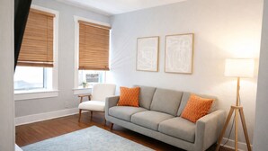 A living room with a grey sofa, two orange pillows, a white chair, a small table, and a floor lamp. - Beautiful 1 Bdrm • Historical • Downtown (Salt Lake City)