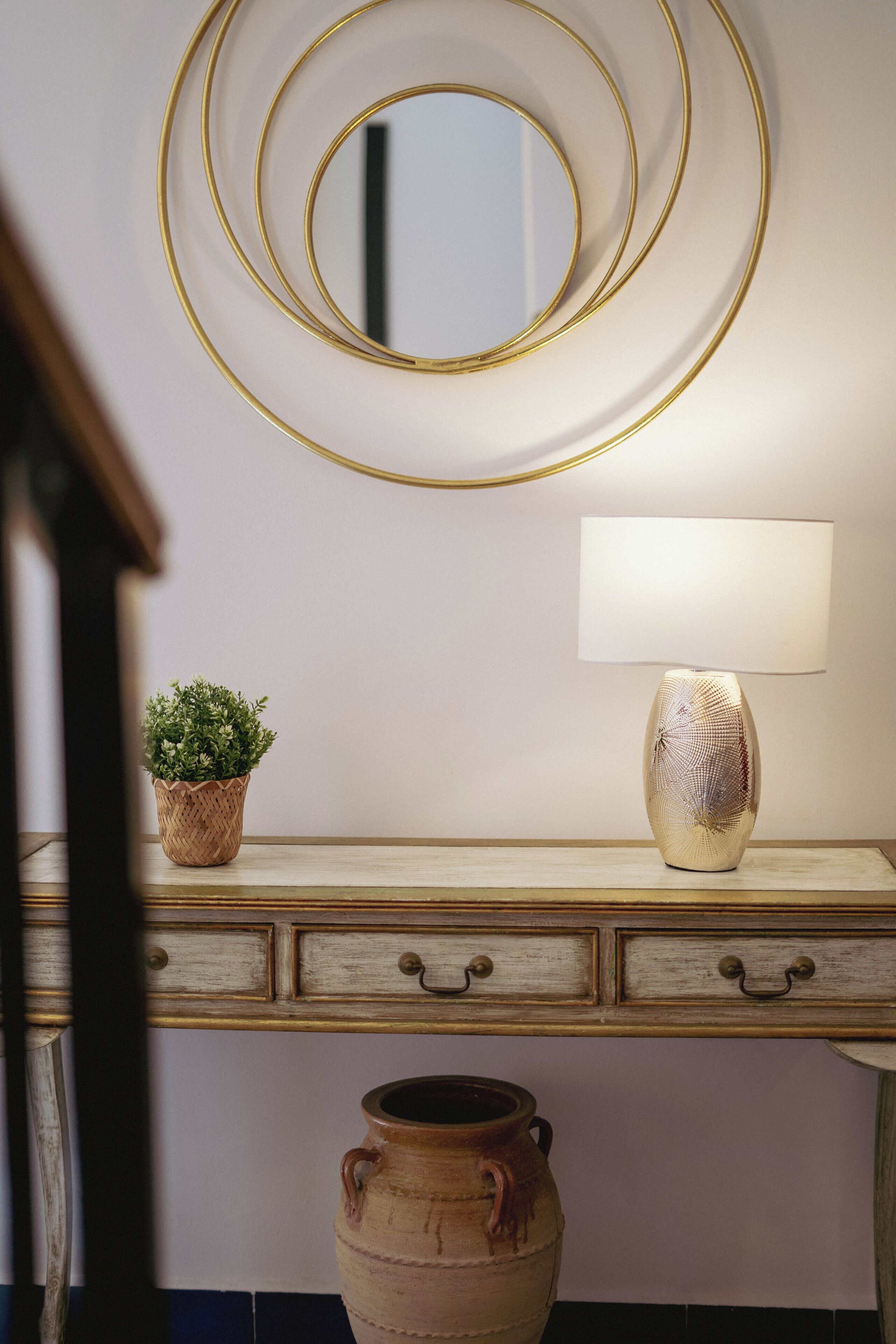 A room with a wooden console table, a mirror with a gold frame, a lamp, a small potted plant, and a large ceramic pot.