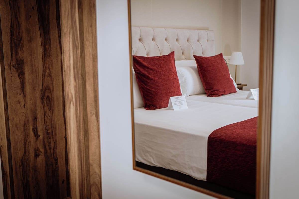 Habitación de hotel con un espejo grande que refleja una cama con almohadones rojos, un cabecero blanco y una mesita de noche con una lámpara.