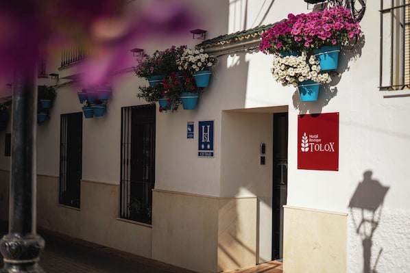A building with hanging flower baskets, a sign for 'Hostal Boutique TOLOX,' and a door labeled 'H.'