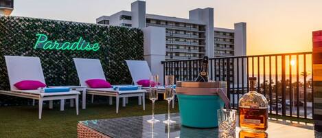 A rooftop lounge with lounge chairs, a neon sign reading 'Paradise,' a cocktail glass, a bottle of liquor, and a teal bucket.