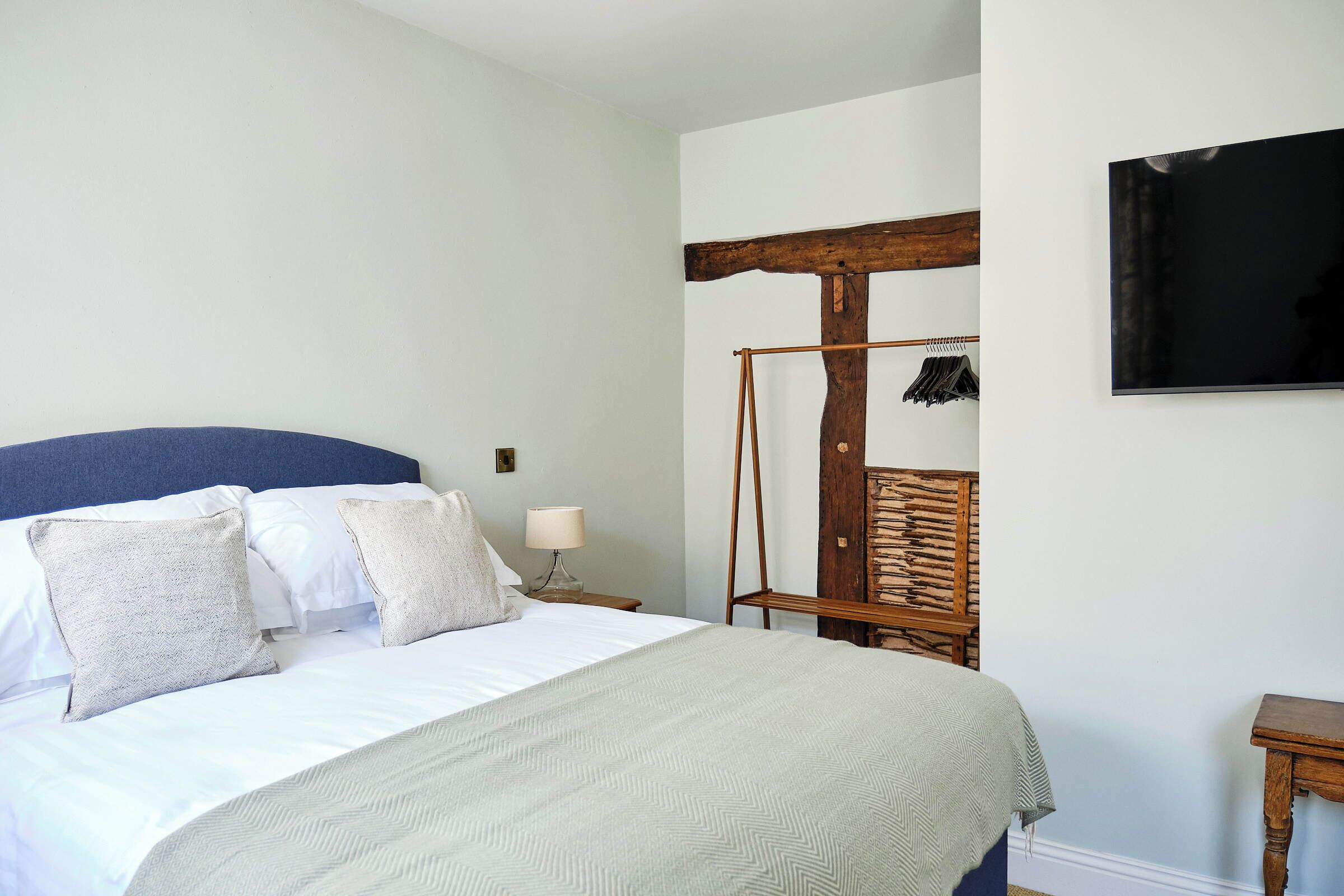 A neatly made bed with a blue headboard, a wooden chair, a small table, and a wall-mounted TV.