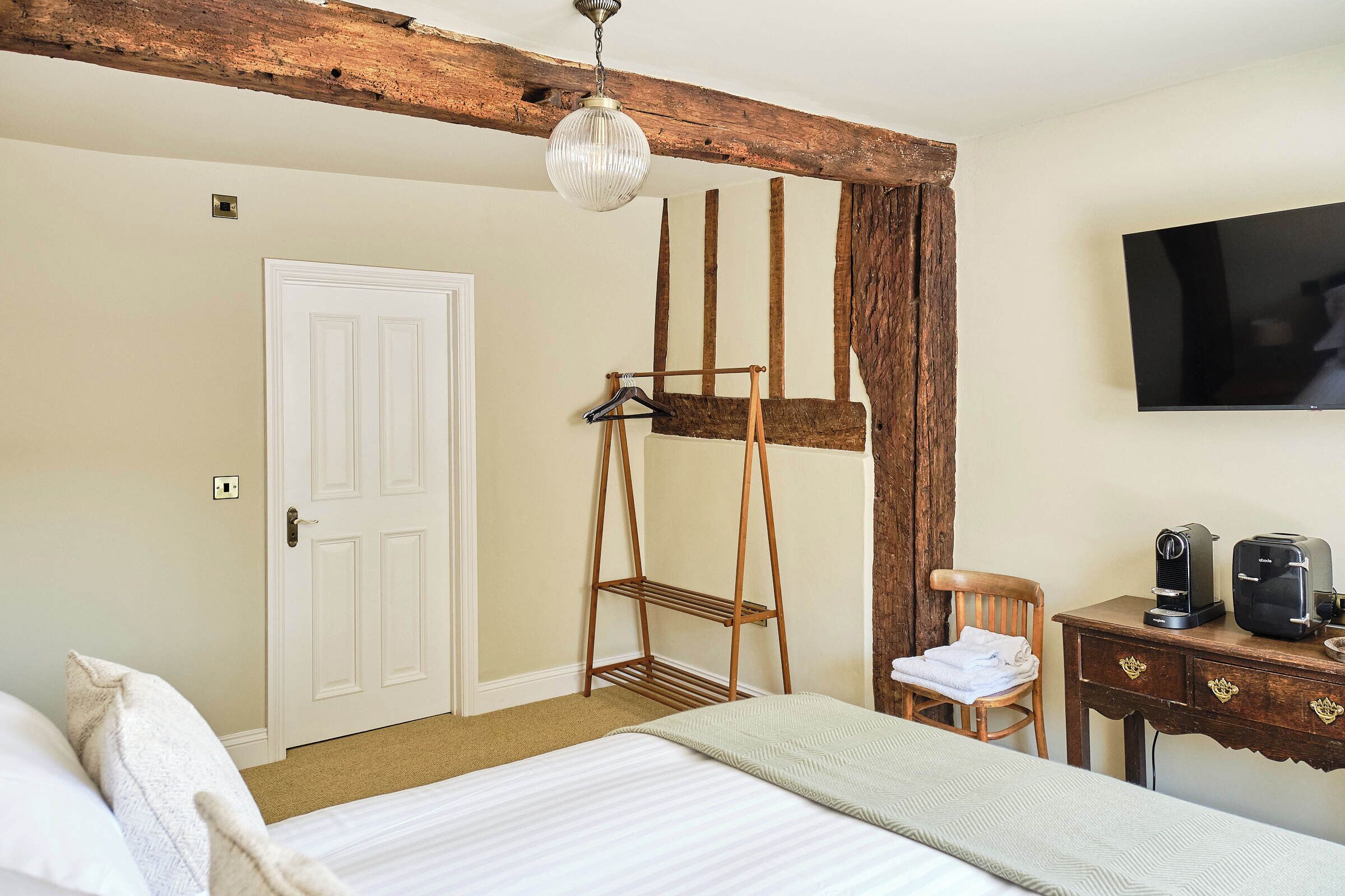 A bedroom with a bed, a wooden chair, a desk with a coffee maker, a TV, and a coat rack.