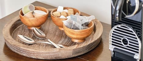 A wooden tray with bowls containing tea bags and sugar cubes, placed on a wooden table next to a coffee machine.