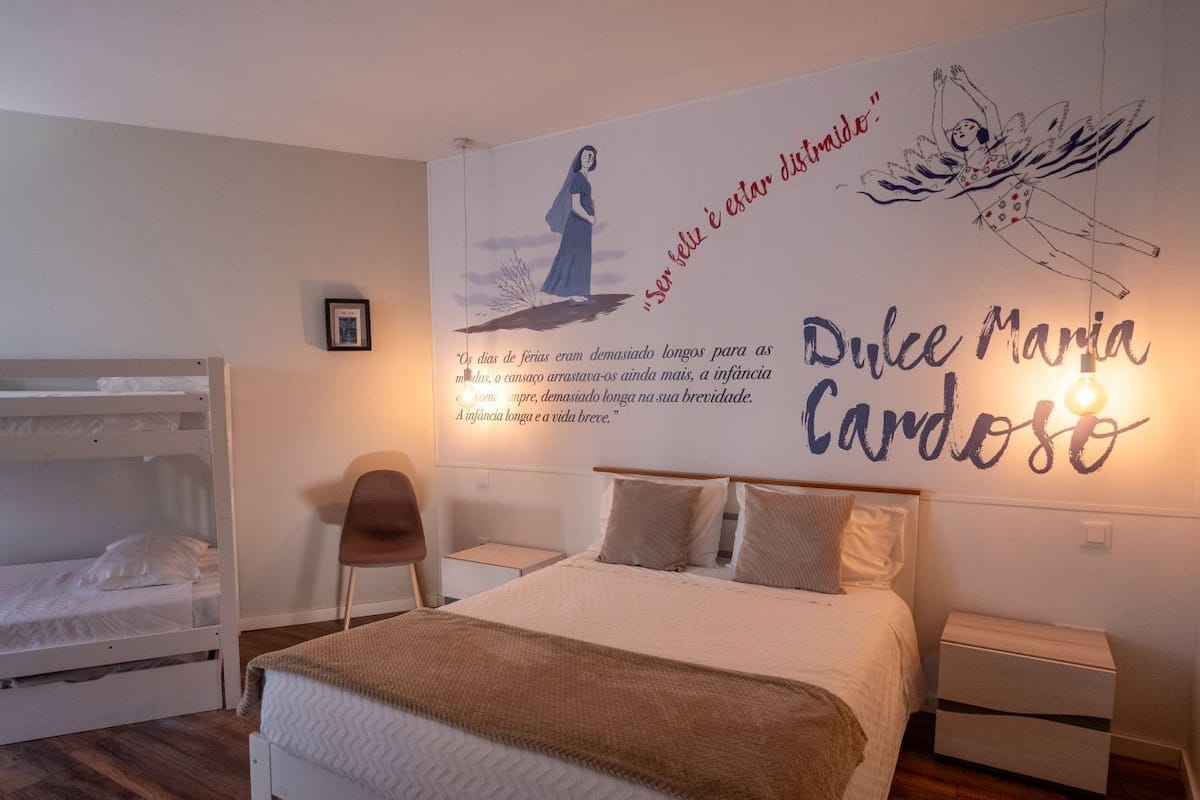 A bedroom with a bunk bed, a single bed, a bedside table, a chair, and a wall with a mural and text.