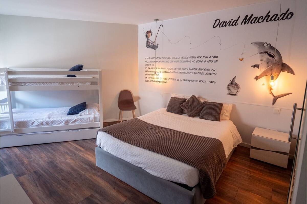 A bedroom with a bunk bed, a single bed, a wall with a David Machado mural, and a small chair.