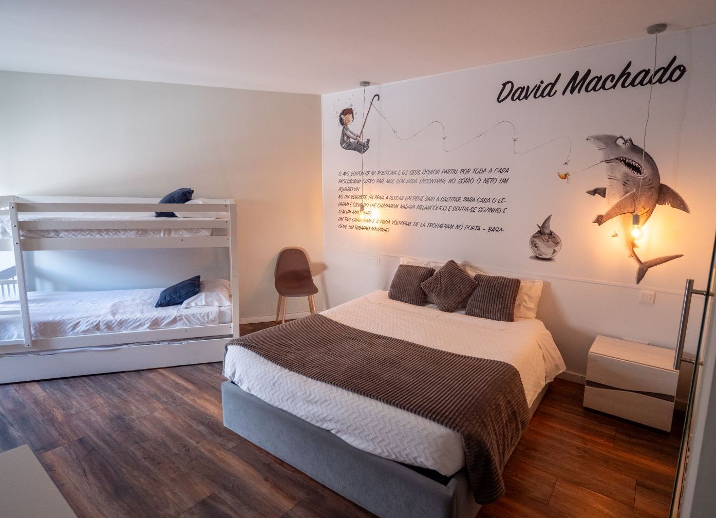 A bedroom with a bunk bed, a single bed, a wall with a David Machado mural, and a small chair.