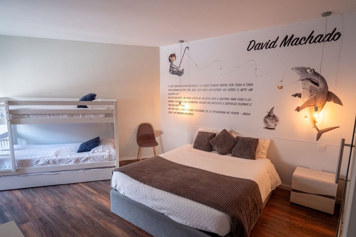 A bedroom with a bunk bed, a single bed, a wall with a David Machado mural, and a small chair.