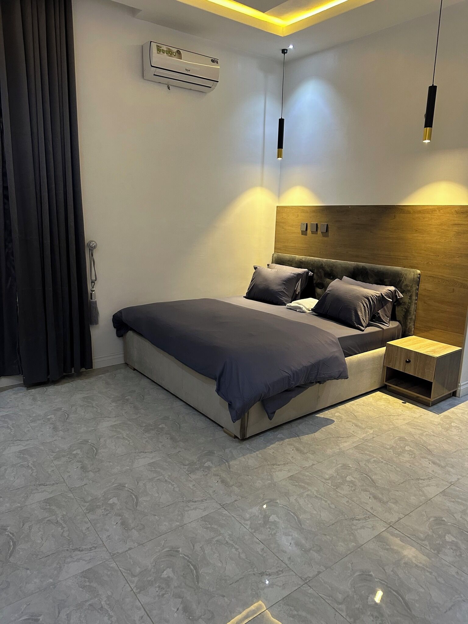 A modern bedroom with a bed, bedside tables, an air conditioning unit, and a ceiling light fixture.