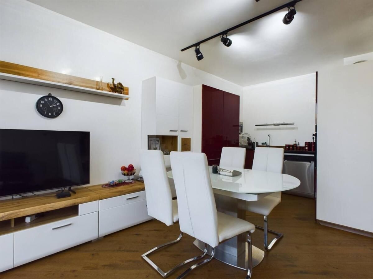Une cuisine moderne avec des meubles blancs, un plan de travail en bois, une table à manger avec des chaises blanches et un téléviseur encastré.