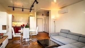 Een moderne woonkamer met een grijze bank, een eethoek met een witte tafel en stoelen, en een keuken op de achtergrond.