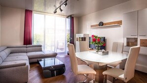 Een moderne woonkamer met een grijze modulaire bank, een glazen salontafel en een eethoek met een witte tafel en vier stoelen. Boven de eettafel staat een tv op een houten plank, en links is er een raam met gordijnen.