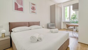 A neatly made bed with white linens and rolled towels, a bedside table with a lamp, a chair, and a desk with a chair. - Bright 1-br apartment in Porta Genova (Milan)