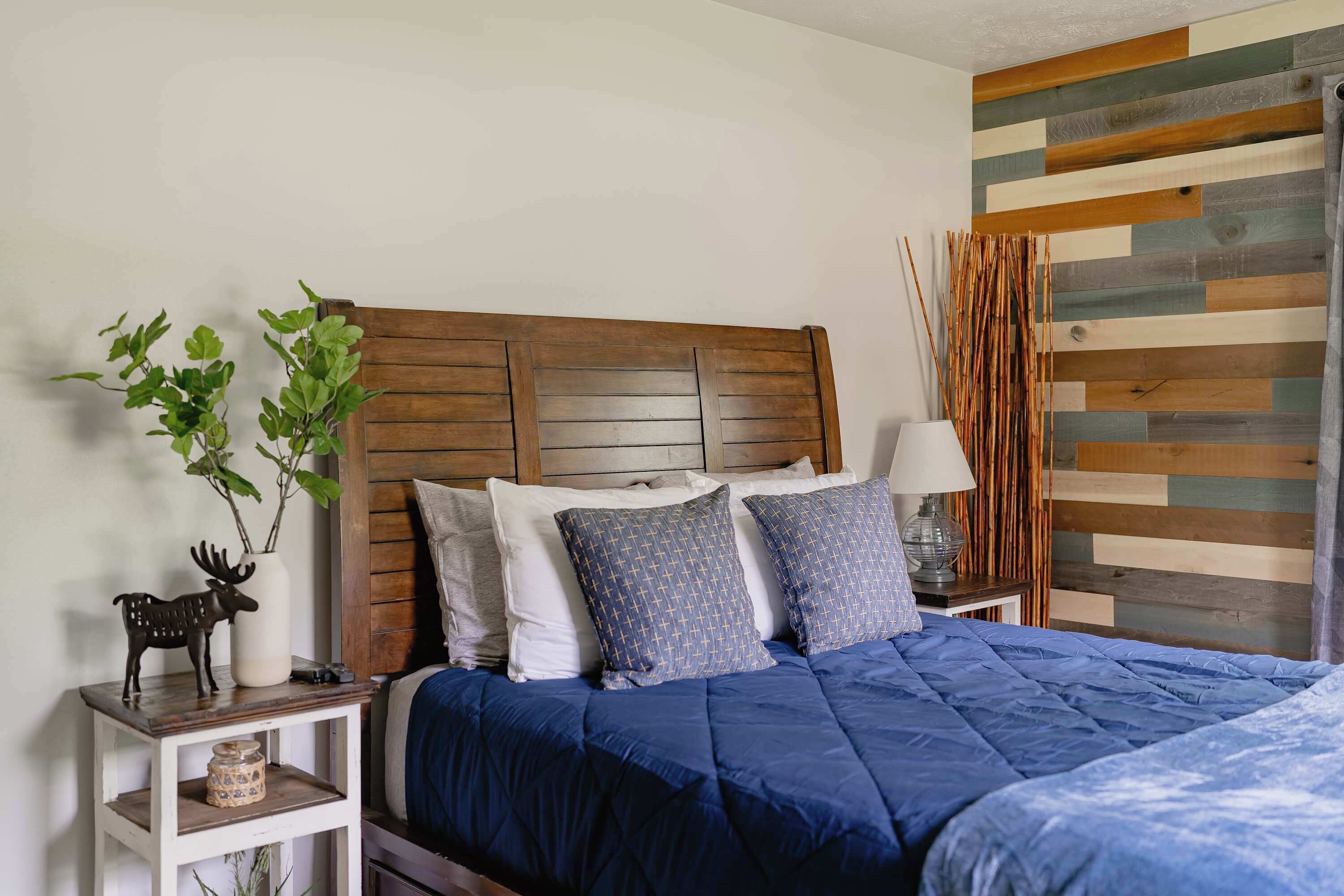 A bedroom with a wooden headboard, a blue bedspread, a nightstand with a lamp, and a decorative plant.